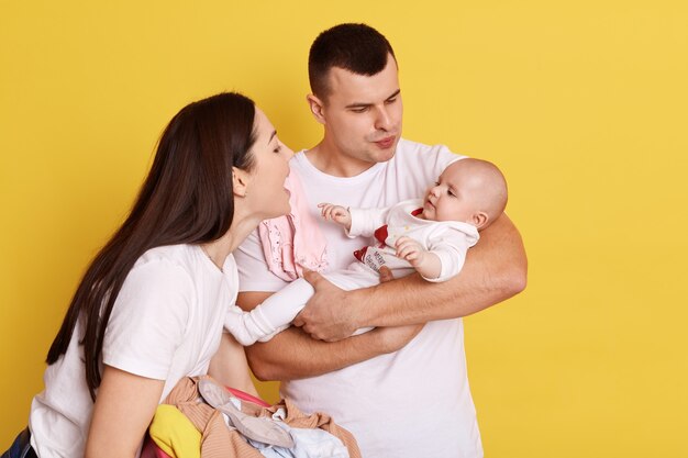 Happy couple embracing and looking at newborn child over yellow background, talking to little daughter with love and smile, parents wearing white t shirts, happy family indoor.