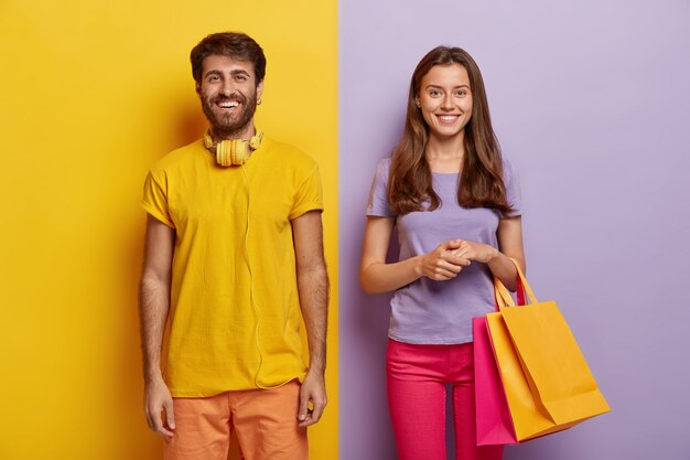 Happy couple enjoy weekend, make purchasing, hold shoppings bags, wears bright outfit, being in high spirit