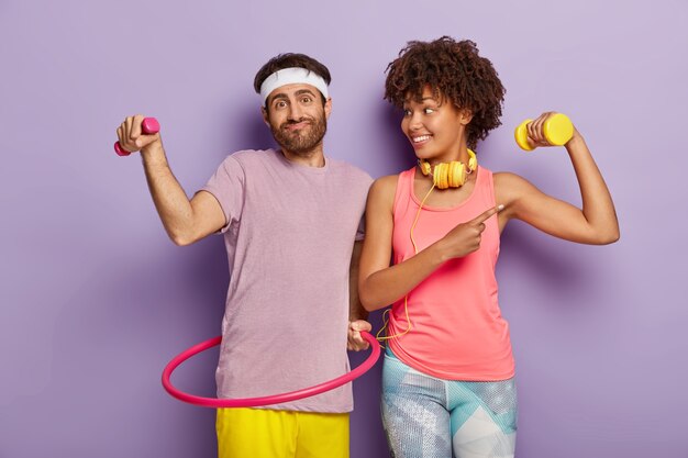 Happy couple have training, like sport, unshaven man holds little dumbbell, exercises with hula hoop, satisfied dark skinned girl shows biceps, trains with weight, listens music in headphones