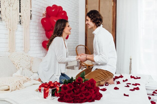 Happy couple holding hands, bouquet of red roses and gift box on the bed