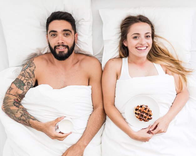 Happy couple lying on bed with cup of coffee and waffles on plate