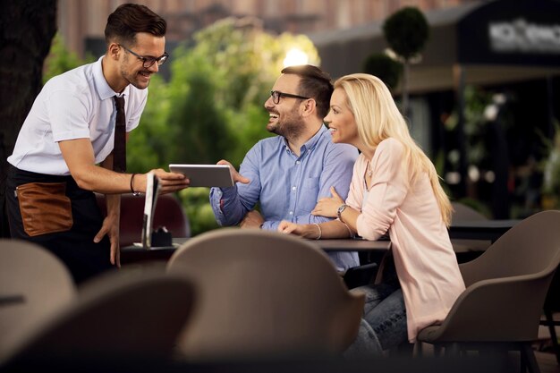 Happy couple making an order in a cafe while waiter is showing them menu on digital table