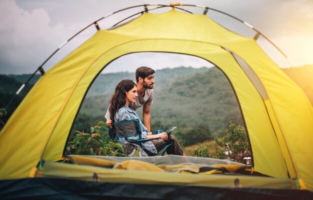 Happy couple man and woman sitting on chair in front of camping tent at campsite in the morning with mountain background