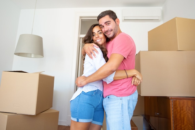 Happy couple moving into new apartment, standing among carton boxes and hugging