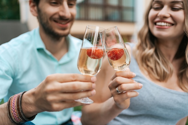 Happy couple portrait clinking two glasses with sparkling wine and strawberries inside with blurred house