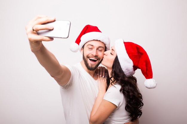 Happy couple in Santa hats waving and taking selfie on smartphone, isolated on white background