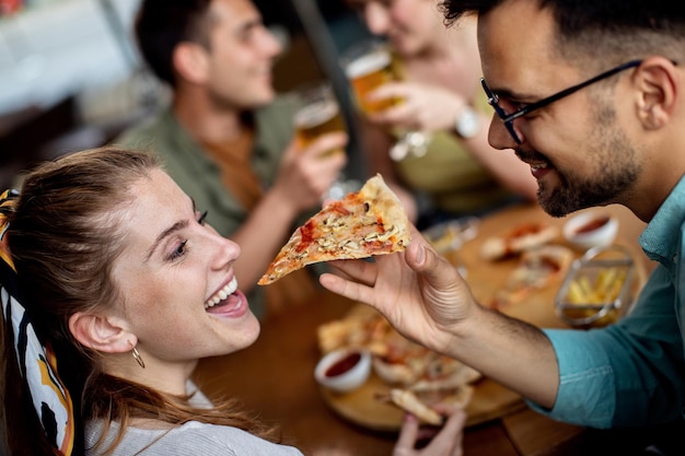 Happy couple sharing slice of pizza while eating with friends in a pub