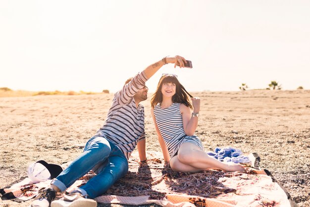 Счастливая пара, сидя на пляже, принимая автопортрет на мобильный телефон