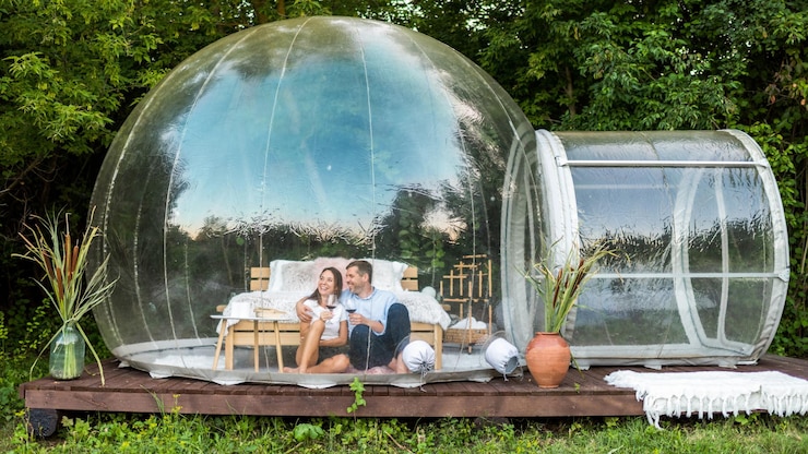 Free Photo a happy couple sitting inside transparent bubble tent at glamping and drinking wine