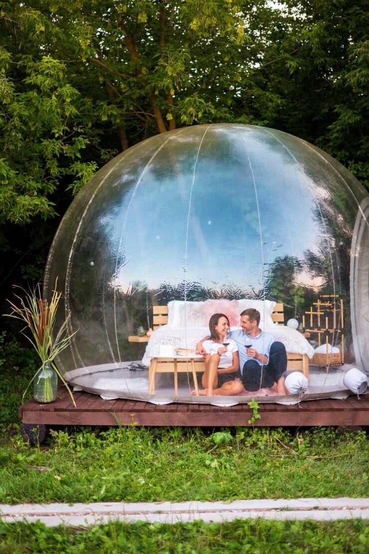 Free Photo a happy couple sitting inside transparent bubble tent at glamping and drinking wine