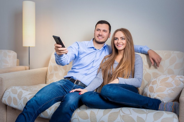 Счастливая пара сидит на диване с пультом дистанционного управления в руках и вместе смотрит телевизор