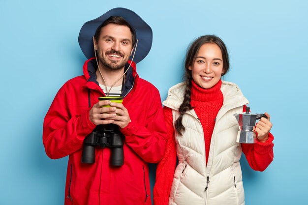 Happy couple spend weekend in nature, drinks coffee, enjoy being on fresh air, use binoculars, dressed in warm casual clothes, stands next to each other over blue wall