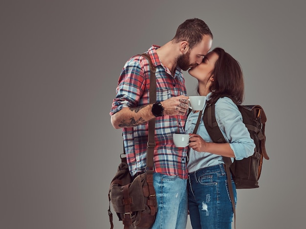 Happy couple of tourists kissing while holding cups of a coffee. Isolated on a gray background.