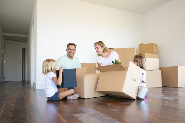 Happy couple and two girls moving into new empty apartment, sitting on floor near open boxes