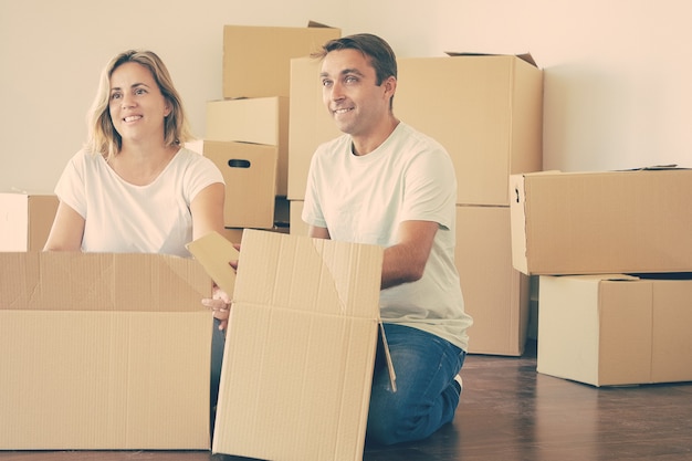 Happy couple unpacking things in new apartment, sitting on floor with open boxes, looking away