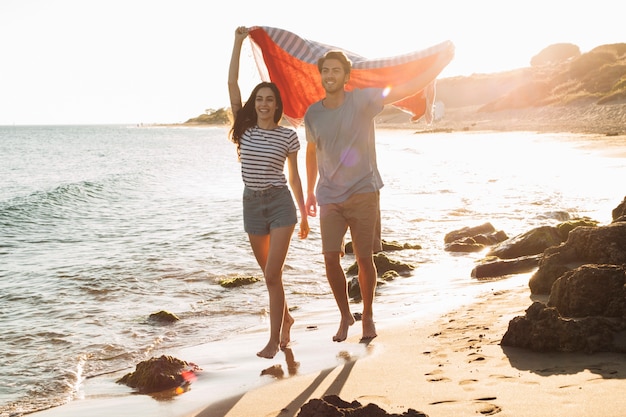 Free photo happy couple walking at the shoreline holding towel in the air