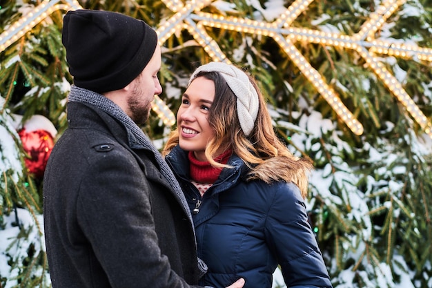 Foto gratuita una coppia felice che indossa abiti caldi si tiene per mano e si guarda l'un l'altro, in piedi vicino a un albero di natale della città, godendosi il tempo trascorso insieme.