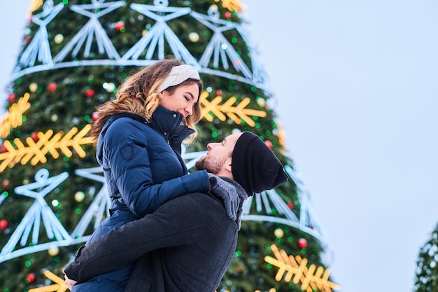 Free photo happy couple wearing warm clothes hold hands and look at each other, standing near a city christmas tree, enjoying spending time together.