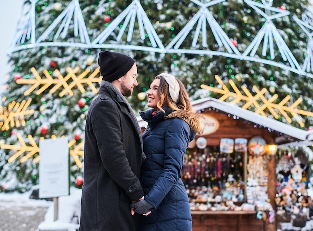 Free photo happy couple wearing warm clothes hold hands and look at each other, standing near a city christmas tree, enjoying spending time together.