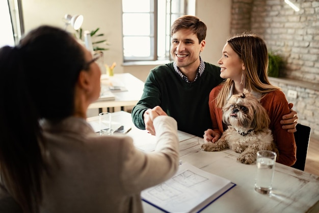 Happy couple with a dog shaking hands with real estate agent in the office