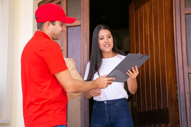 Happy courier showing order sheet to client and holding clipboard. Professional deliveryman carrying paper bag. Woman receiving order at home. Food delivery service and online shopping concept