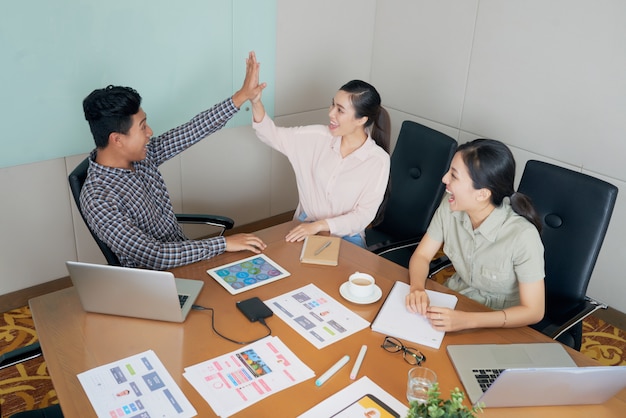 Free photo happy creative asian colleagues sitting at desk in office and doing high-five