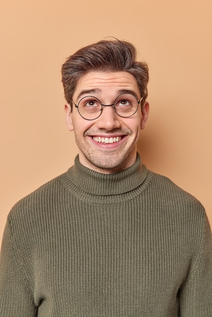 Happy curious man looks with interest above smiles joyfully hears strange noise upstairs dressed casually poses over beige studio wall. Satisfied glad guy focused up smiles toothily wears spectacles