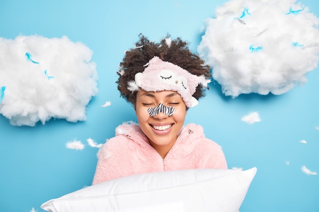 Happy curly Afro American woman gets rid of black dotes on nose using special patch wants to have flawless clean skin smiles gently dressed in pajama holds pillow poses around flying feathers