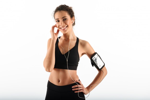 Happy curly brunette fitness woman holding arm on hip and listening music