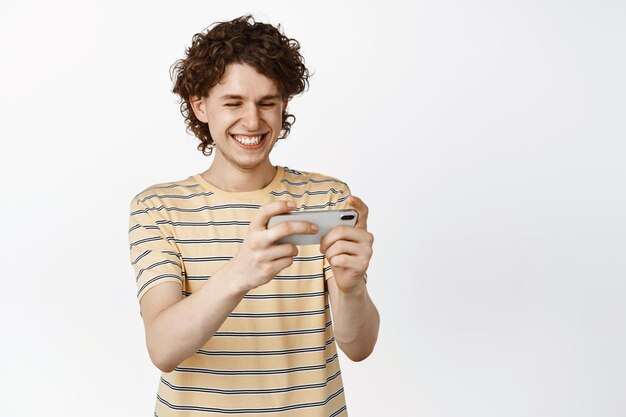 Happy curly guy playing video game on mobile phone watching video on smartphone screen and smiling standing over white background