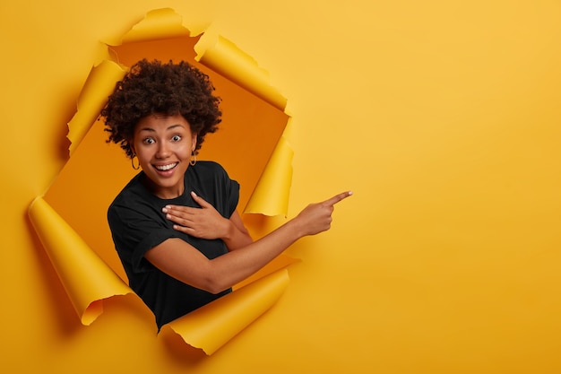 Free photo happy curly haired african american woman laughs positively, points aside on copy space, wears black t shirt