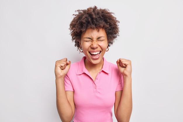 Счастливая кудрявая афро-американка накачивает кулаком и радуется положительным результатам в повседневной розовой футболке.