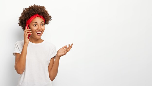 Happy curly haired girl enjoys pleasant telephone conversation, gestures with palm, uses mobile phone, looks aside with glad expression