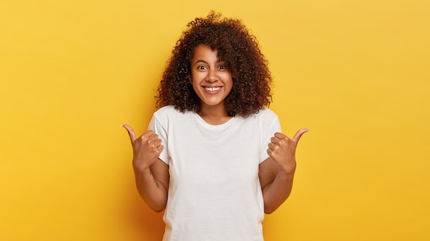 Happy curly haired girl makes thumbs up sign, demonstrates support and respect to someone, smiles pleasantly , achieves desirable goal, wears white t shirt, isolated on yellow wall