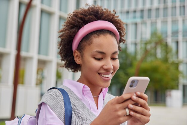 ストリートの服を着た幸せな巻き毛の笑顔の10代の少女は、大学に行く途中でリュックサックを運びますソーシャルネットワークで携帯電話のチェックメールを使用して個人のブログでマルチメディアを共有します