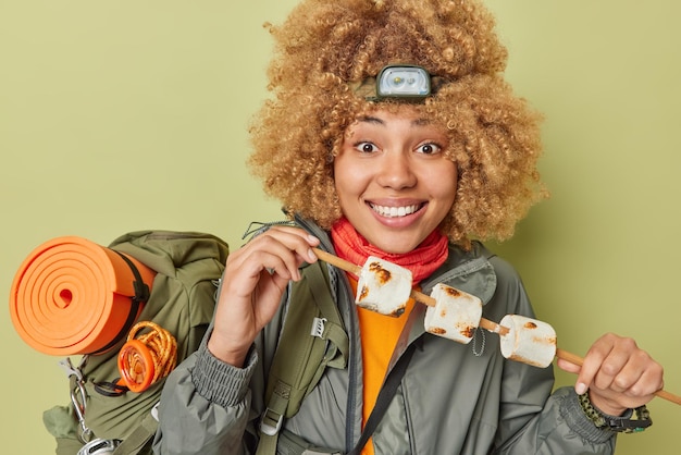 Free photo happy curly haired woman camper holds wooden stick with roasted marshmallow wears headlamp and windbreaker carries rucksack isolated over green background young tourist has picnic during weekend