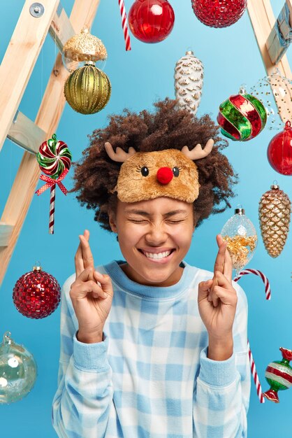 Happy curly haired woman crosses fingers and makes wish on New Year night wears reindeer sleepmask and pajama poses against blue background with toys for decoration around. Holidays concept.