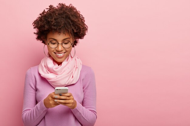 Happy curly haired woman dressed in casual clothes, smiles happily and delighted, wears big earrings, turtleneck with silk scarf