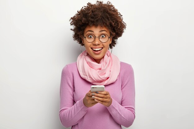 Happy curly haired woman holds modern cellular, chats online, smiles joyfully, wears transparent glasses and casual violet jumper