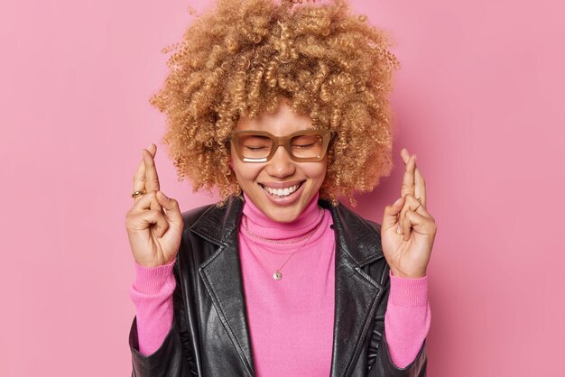 Happy curly haired woman makes hopeful please gesture smiles gladfully keeps eyes closed wears spectacles turtleneck and leather jacket isolated over pink background makes wish prays about something