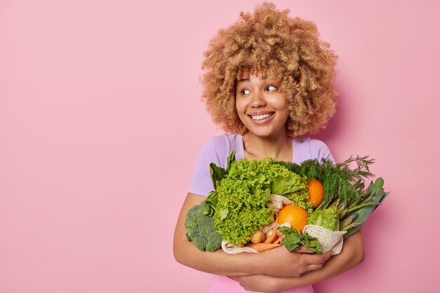 Happy curly haired young woman embraces bouquet of fresh vegetables picked up from own garden smiles broadly dressed in casual t shirt isolated over pink background with copy space for promno