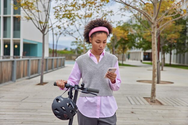 Happy curly millennial girl in her twenties rides kick electric scooter in urban park and checks phone