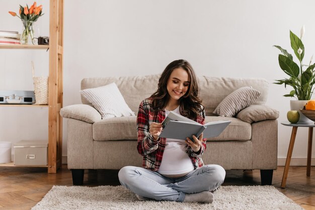Счастливая кудрявая беременная женщина в клетчатой рубашке и джинсах читает книгу Привлекательная брюнетка отдыхает на мягком ковре возле бежевого дивана