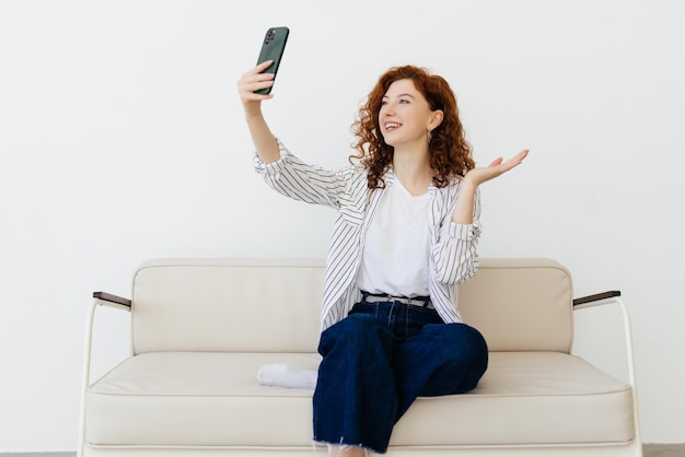 Happy curly redhaired woman resting at home on leather sofa talking chatting via video call using app on smartphone has conversation with family greeting friends waving hand