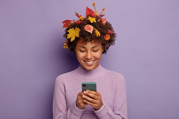 행복한 곱슬 여자는 휴대 전화를 통해 친구와 채팅하고, 부드러운 미소를 지으며, 머리에 노란 잎이 있습니다.