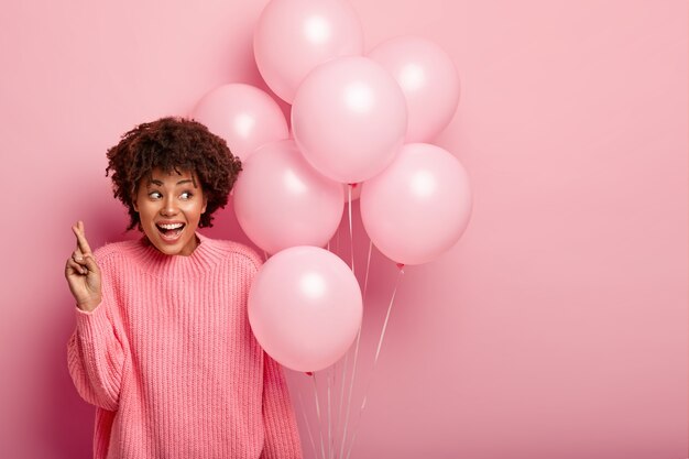 Happy curly woman in oversized jumper, keeps fingers crossed for good luck, holds helium balloons