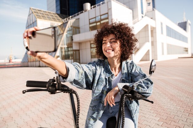屋外の現代的なバイクに座って幸せな巻き毛の女性