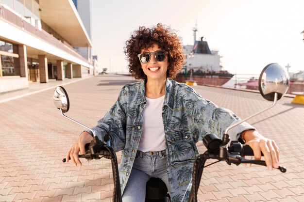 Happy curly woman in sunglasses rides on modern motorbike