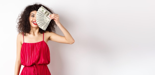 Free photo happy curly woman winning money holding dollar bills on half of face smiling excited standing in red