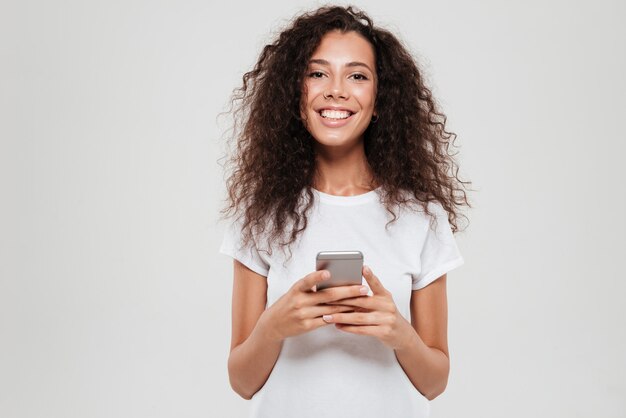 Happy curly woman with smartphone in hands looking at the camera over gray background